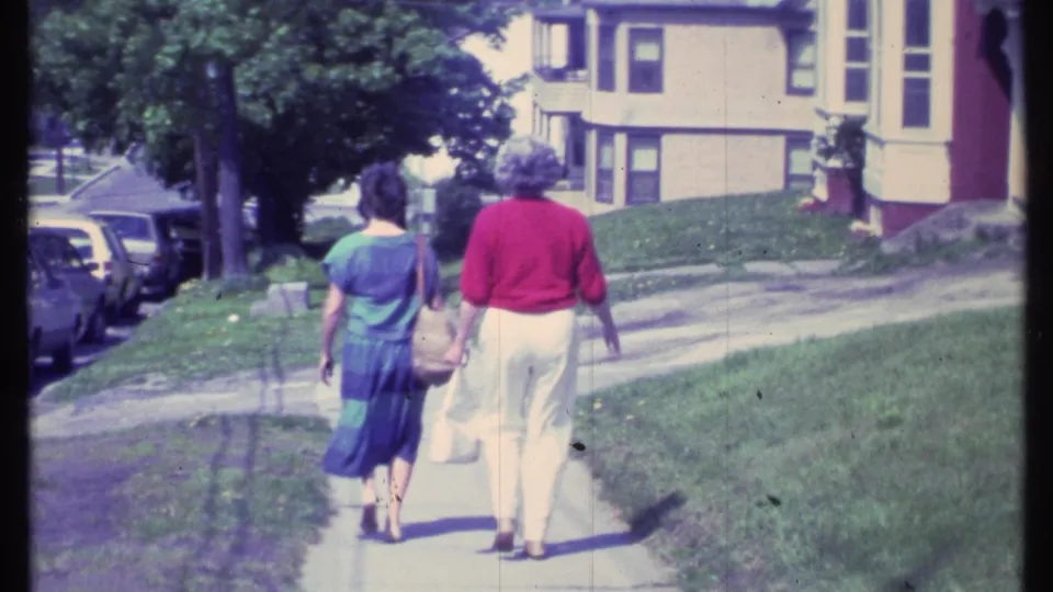 The pair continues down the sidewalk in the Vermont 1984 home-movie shot, passing lawns, mature trees, parked cars, and a house facade.