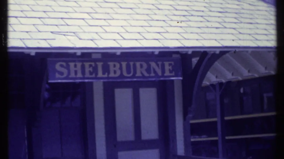 A station sign reading SHELBURNE appears under a canopy near a platform, with the side of a train visible in the 8mm frame.