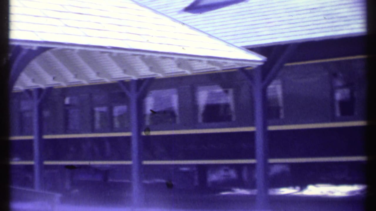 1984 Vermont 8mm frame showing a dark passenger rail car beside a covered platform, with curtained windows and a purple film cast.