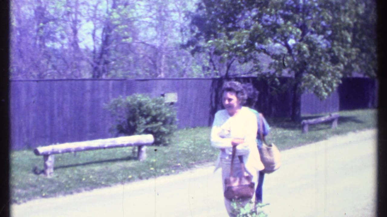 In Vermont 1984 home-movie footage, an older woman with a handbag stands by a paved park path near a wooden bench and dark fence.