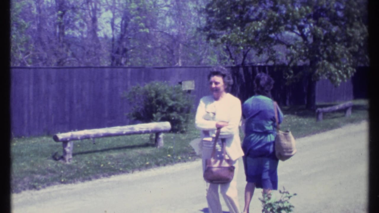 Two older women with handbags stand on a paved path beside benches, fence, trees, and lawn.