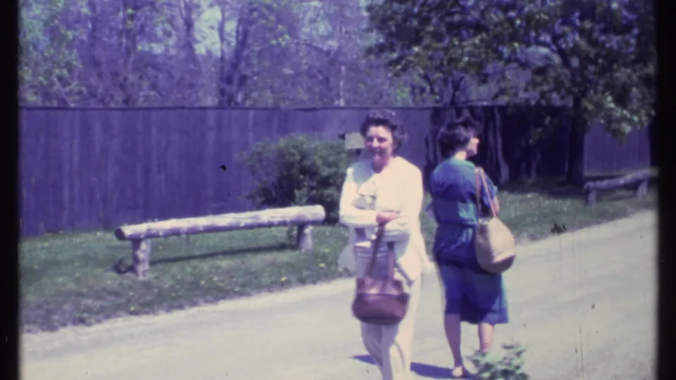 Two older women walk along a paved path in 1984 Vermont footage, passing a wooden bench with a dark fence and trees in the background.