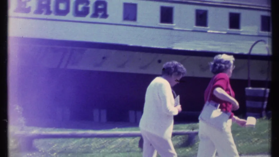 Two older women walk near a large white building with partial visible signage, with a bench, lawn, and trees in the surrounding area.