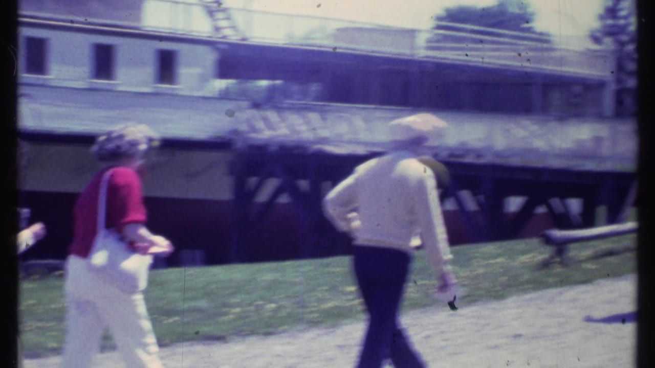 A woman in red and a man in a light sweater walk near a docked riverboat and pier.