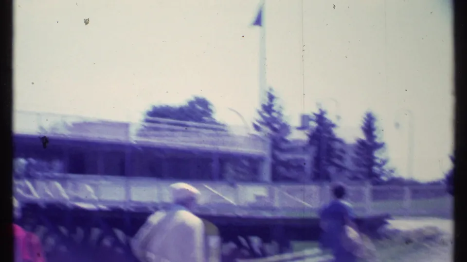 The home-movie frame looks across a Vermont riverside path toward visitors, trees, and the dock area beside the riverboat.