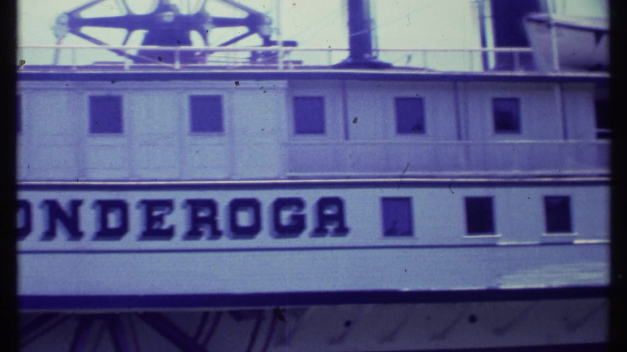 Side of the vessel with large Ticonderoga lettering, square windows, railings, smokestack, and paddlewheel structure above.