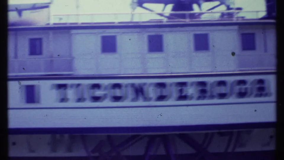 1984 8mm frame of the Ticonderoga hull lettering in motion blur, with a row of windows, deck railings, and paddlewheel structure above.