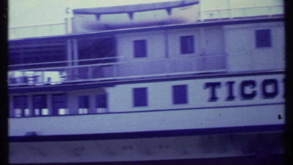 8mm frame from Vermont showing a cropped side section of the Ticonderoga with partial name lettering, rows of windows, railings, and upper-deck boat equipment.