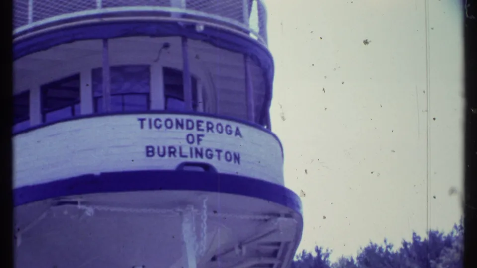 Closer 8mm frame of the Ticonderoga of Burlington hull lettering with deck railings and trees in the background.