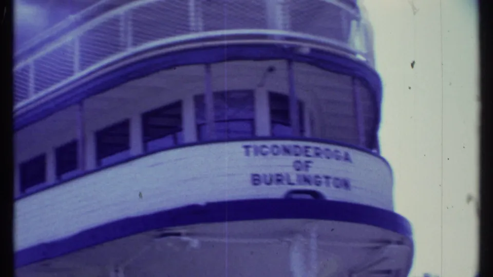 Angled handheld view of the Ticonderoga of Burlington bow, with windows, railing, lettering, and pale sky visible.