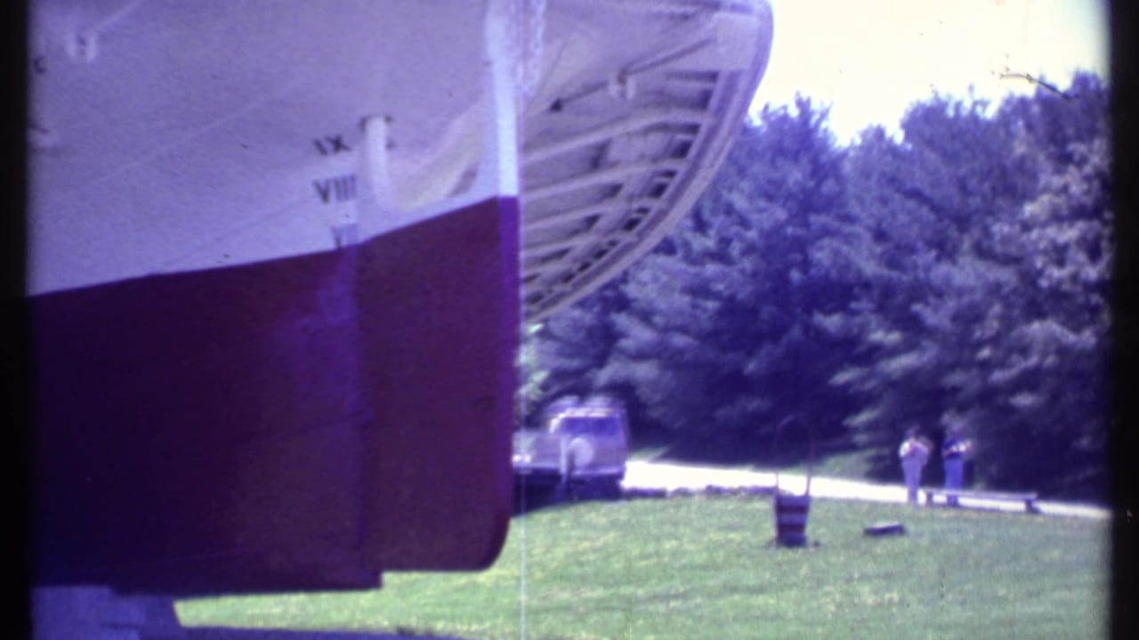 Boat stern area beside grass and trees, with a parked vehicle and distant people visible.