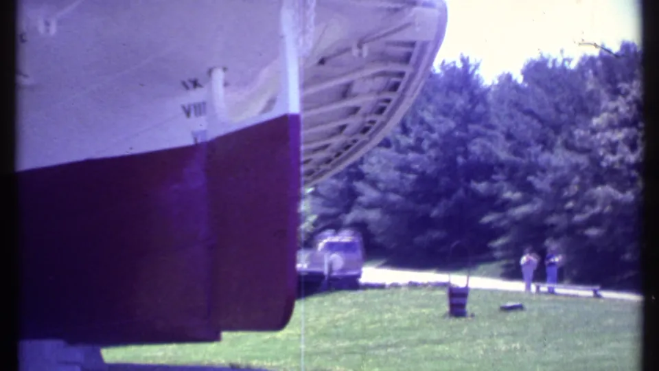The docked vessel's stern and hull are seen near a grassy lakeshore with trees, a vehicle, and distant visitors.