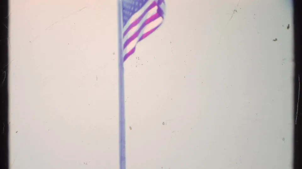 1984 Vermont 8mm frame with a U.S. flag fluttering near the top of a tall pole against a washed pale sky.
