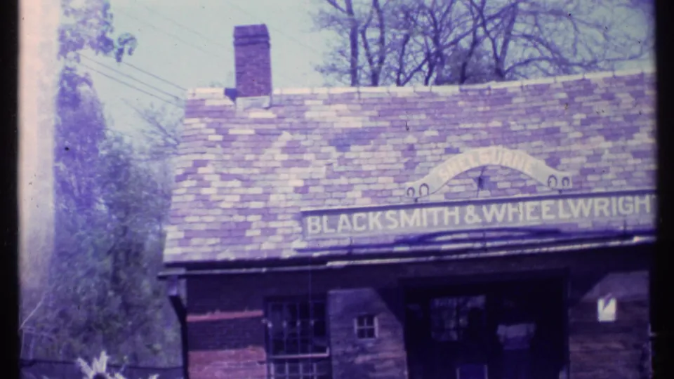 1984 Vermont 8mm frame showing a rustic shopfront with a slate-like roof and BLACKSMITH & WHEELWRIGHT sign.