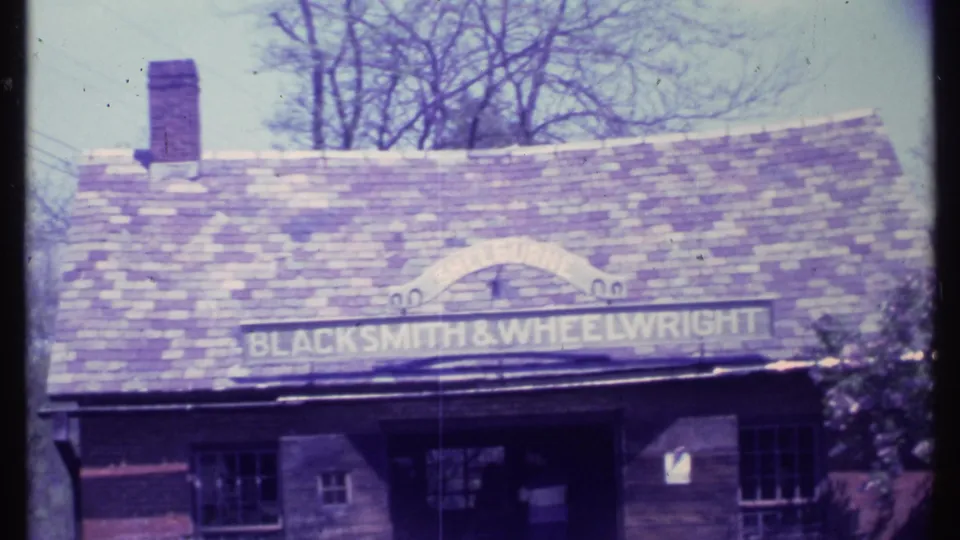 1984 Vermont 8mm frame of the BLACKSMITH & WHEELWRIGHT building exterior with chimney, roof, windows, and bare trees behind it.