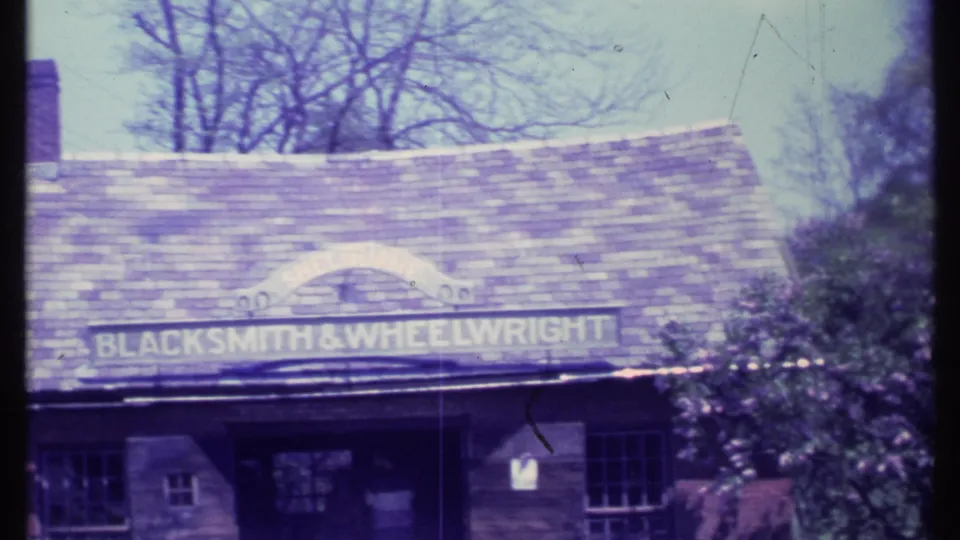 1984 8mm frame of the Blacksmith and Wheelwright shop exterior in Vermont, showing the roof, arched sign, doorway, window, and shrubs.