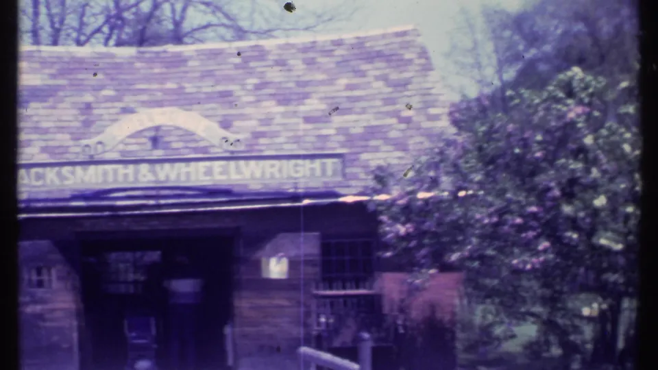 Visitor-shot 1984 Vermont frame showing the Blacksmith and Wheelwright shop sign, entrance area, window, roofline, and surrounding shrubs.