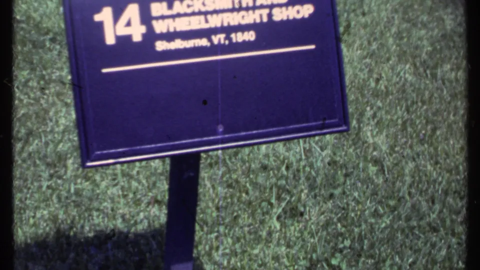 1984 Vermont frame focused on a purple site marker for Blacksmith and Wheelwright Shop, number 14, with grass and part of the building behind it.