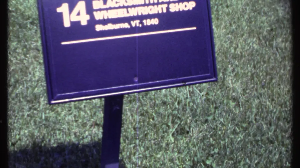 8mm frame from Vermont showing the number 14 Blacksmith and Wheelwright Shop sign in grass, with the preserved building facade behind it.