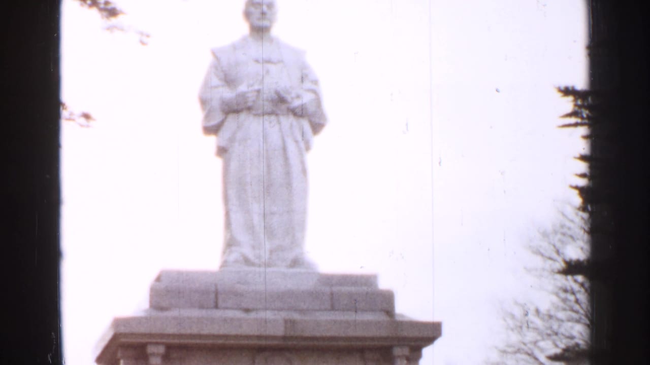 Stone statue in robes on a pedestal against a bright sky with trees at the edge.