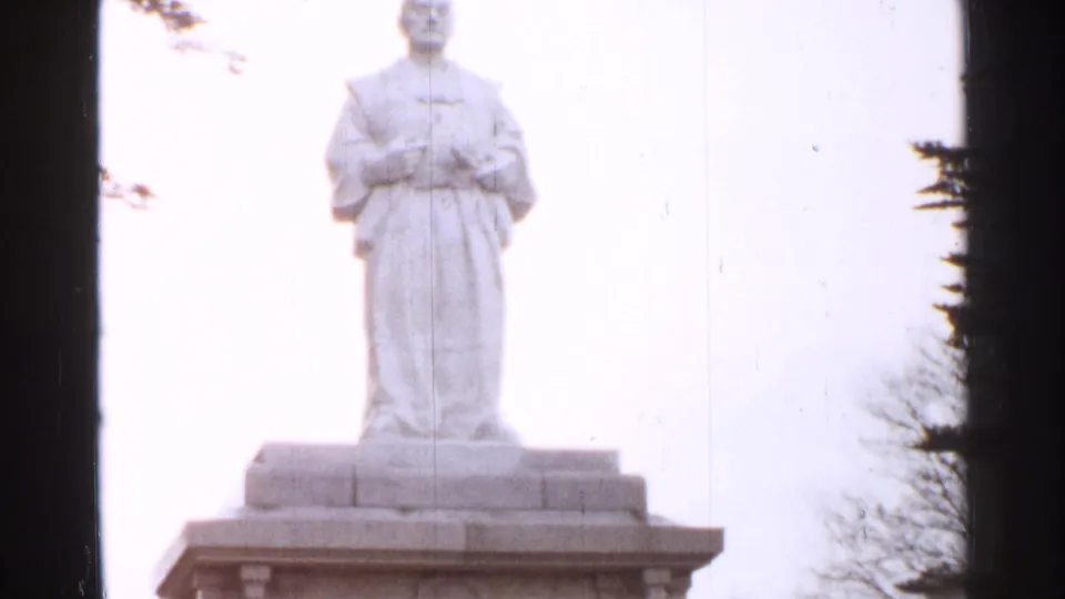 A robed stone statue stands on a pedestal in Japan, filmed in soft 8mm color against a pale sky with trees at the margins.