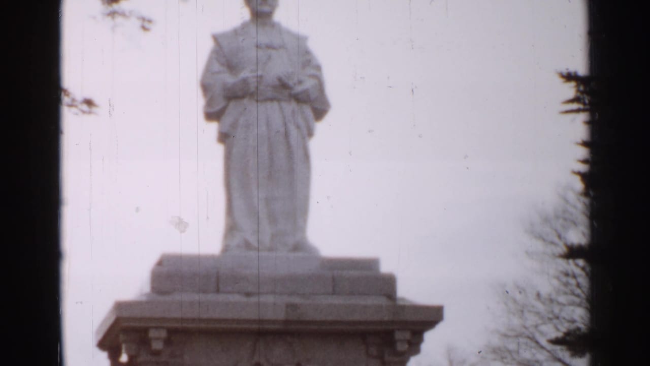 The 1952 Japan footage holds on the robed stone statue and its broad pedestal, with bare branches and dark frame edges visible.