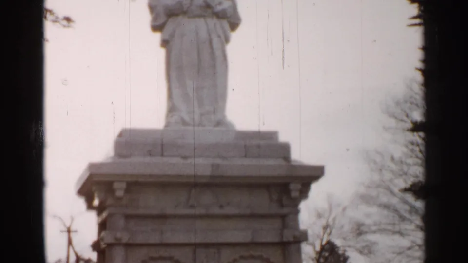 1952 Japan 8mm frame showing the lower part of a white statue on an ornate stone pedestal with bare trees nearby.