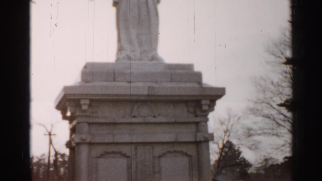 The camera holds on a weathered white statue pedestal in Japan, with bare trees and utility poles visible in the background.