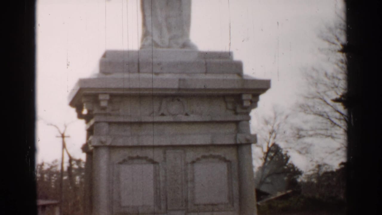 A closer 8mm view of the statue pedestal shows carved panels, lower steps, bare branches, and a muted outdoor setting.