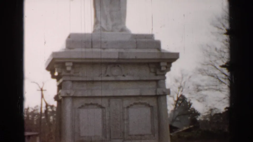 A closer 8mm view of the statue pedestal shows carved panels, lower steps, bare branches, and a muted outdoor setting.