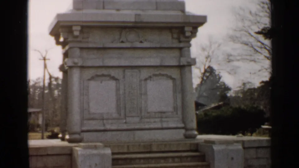 1952 Japan home-movie frame with a centered white stone pedestal, carved side panels, low steps, trees, and utility poles in view.