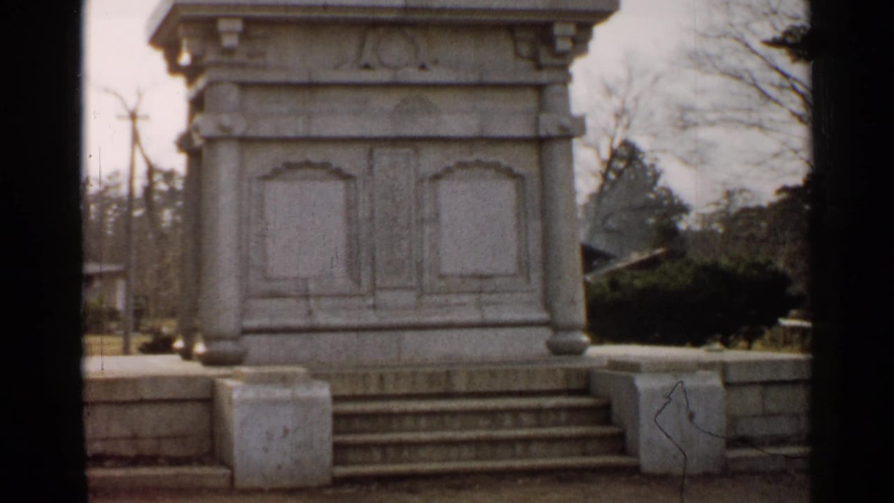1952 8mm Japan frame showing the stone pedestal, shallow steps, low surrounding wall, grass, trees, and distant utility poles.