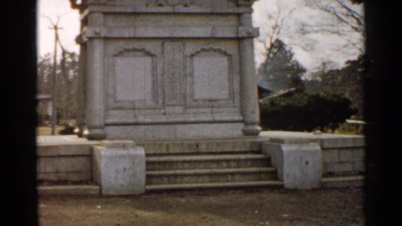 1952 Japan 8mm frame with a fuller view of the white stone pedestal, steps, low wall, shrubs, bare trees, and cloudy outdoor grounds.