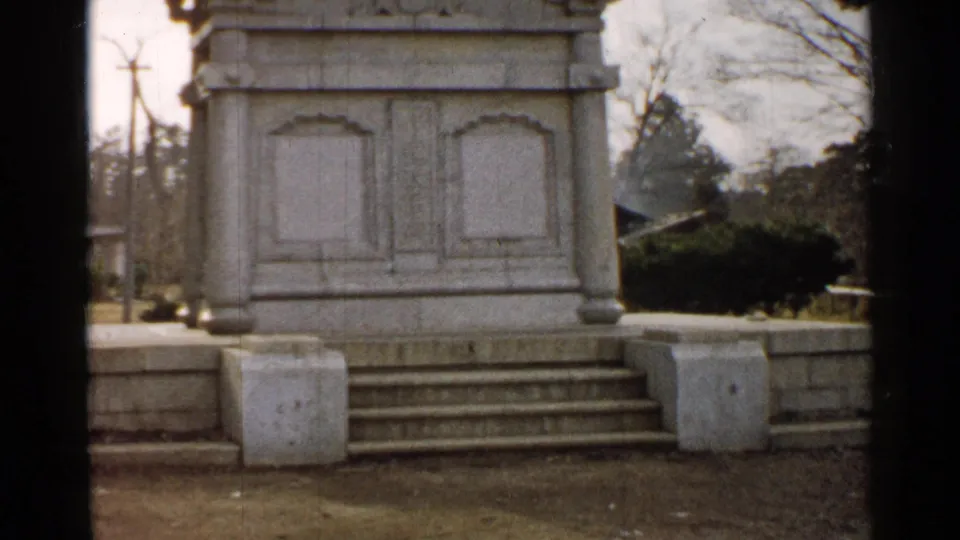 1952 Japan 8mm frame with a fuller view of the white stone pedestal, steps, low wall, shrubs, bare trees, and cloudy outdoor grounds.