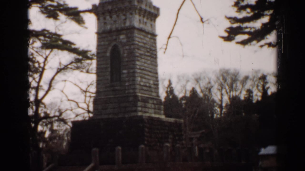 1952 Japan 8mm frame showing a tall weathered stone monument partly framed by trees under a pale sky.