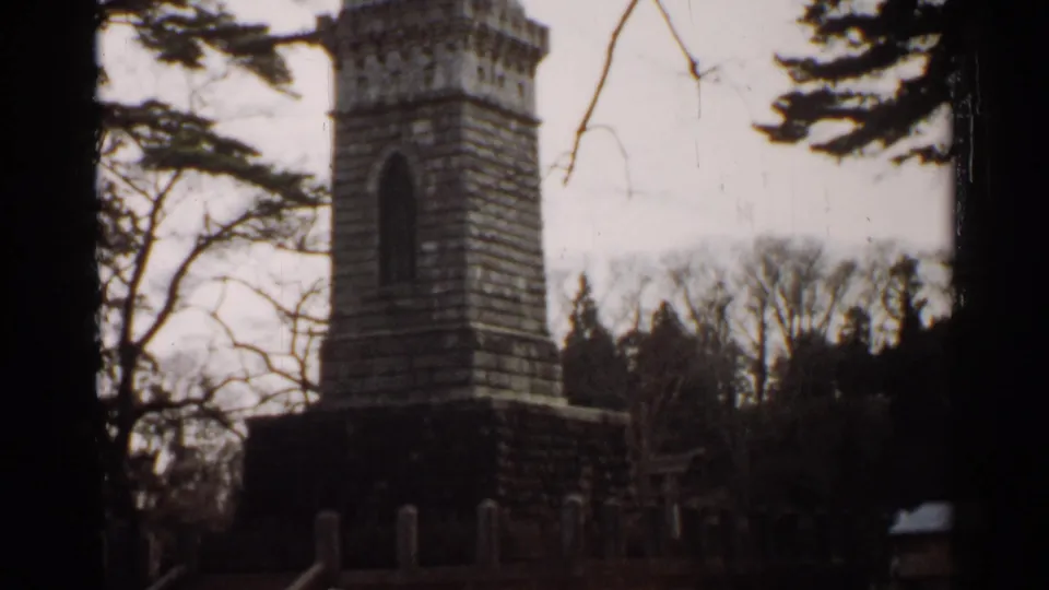 1952 Japan 8mm frame showing a tall weathered stone monument partly framed by trees under a pale sky.