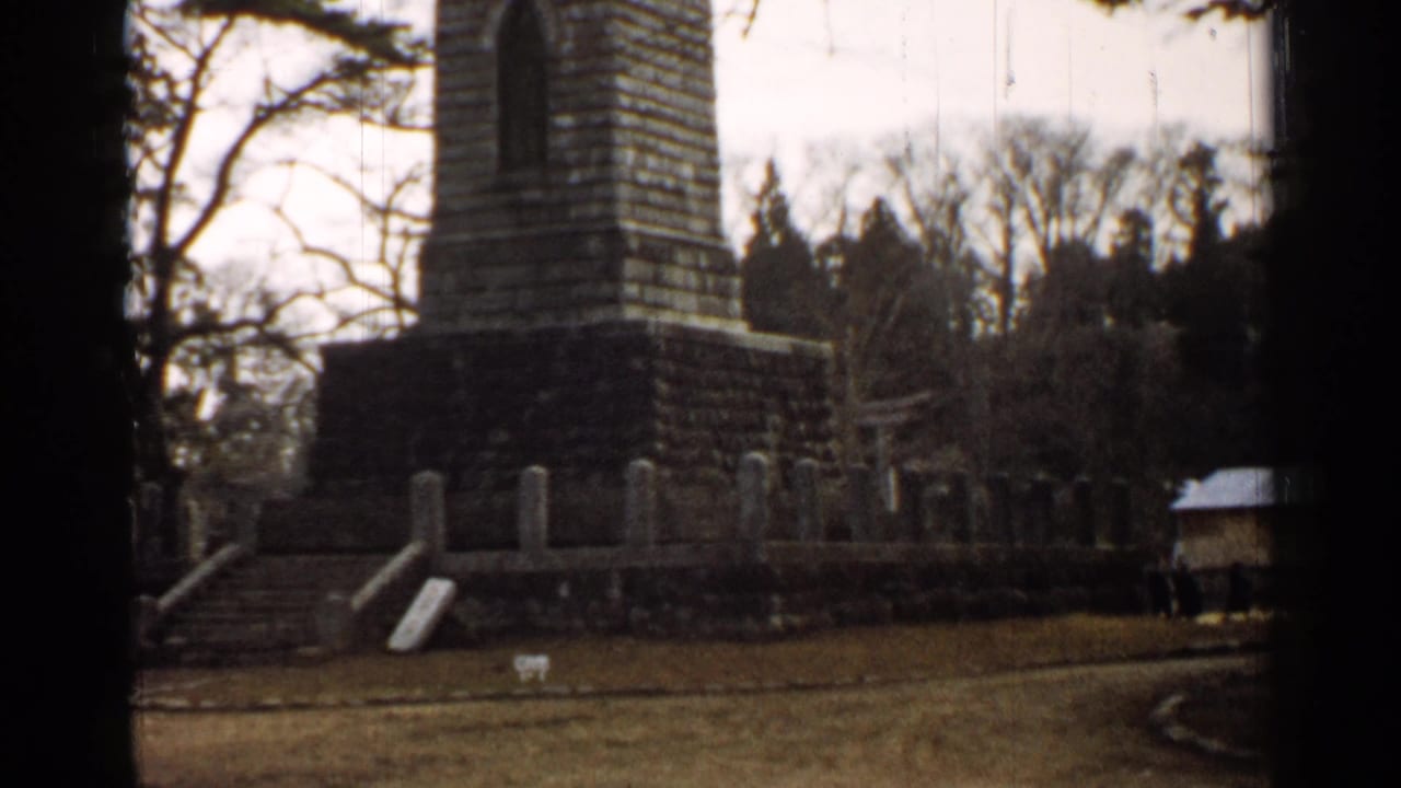 1952 Japan 8mm frame with the monument base, steps, low wall, grass foreground, and wooded backdrop in soft focus.
