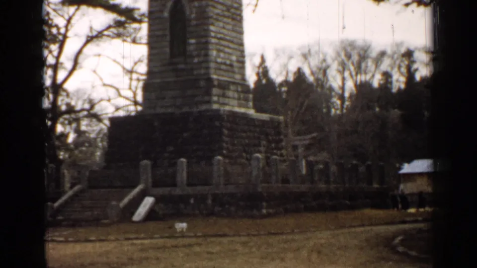 1952 Japan 8mm frame with the monument base, steps, low wall, grass foreground, and wooded backdrop in soft focus.