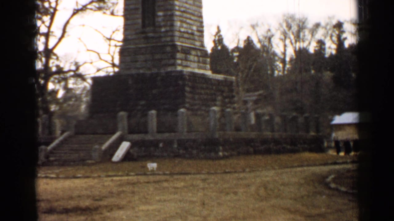 1952 Japan 8mm frame showing the lower monument, grassy field, steps, wall markers, trees, and a small structure at right.