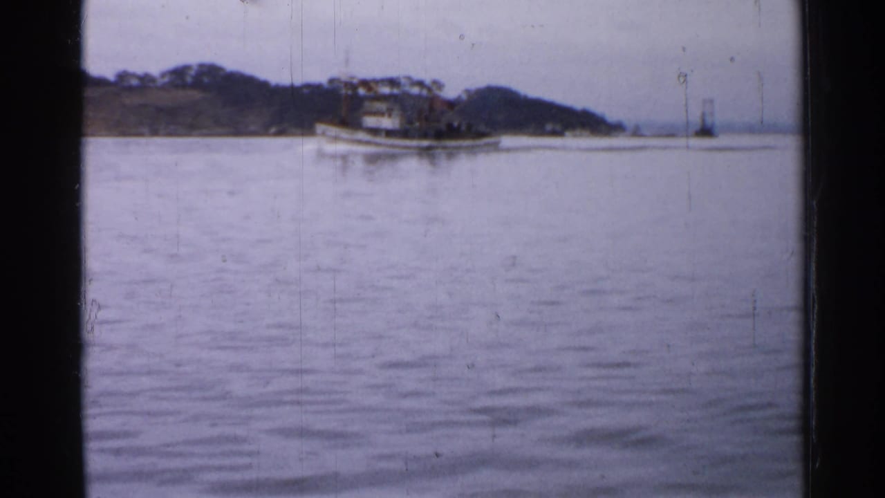 A white tugboat crosses calm water near a low wooded shoreline in Japan, captured on faded 8mm color film.