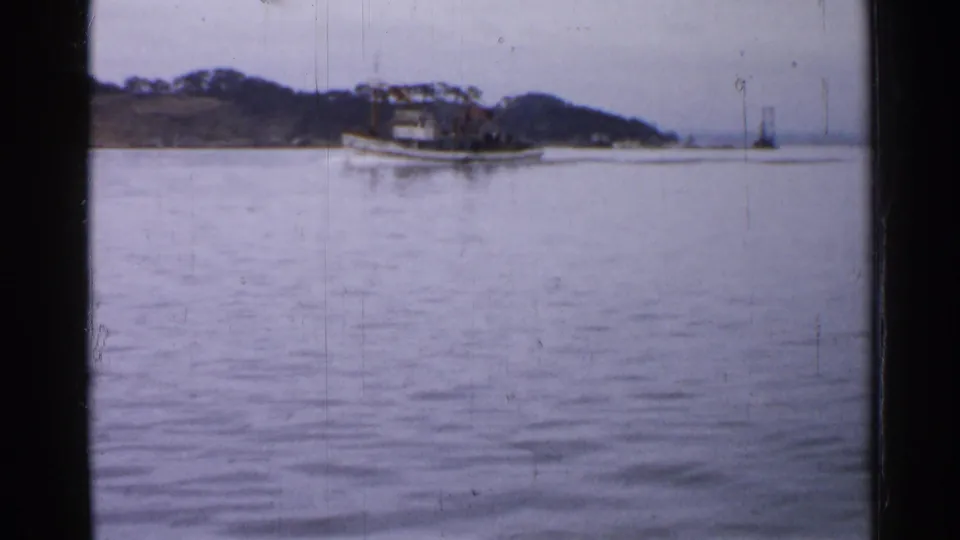 A white tugboat crosses calm water near a low wooded shoreline in Japan, captured on faded 8mm color film.