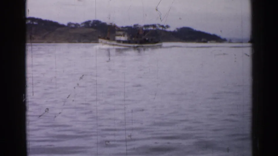 A white tugboat sits farther across calm water near a wooded bank, framed by vintage 8mm scratches and fading.
