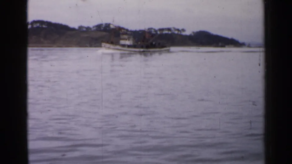1952 Japan 8mm frame showing a small boat moving across calm water with wooded shoreline and hazy hills behind it.