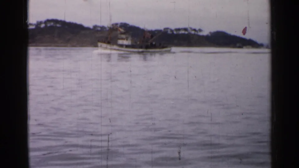 Small boat on calm Japanese waters in 1952, seen from a distant 8mm home-movie viewpoint with tree-lined shore behind.