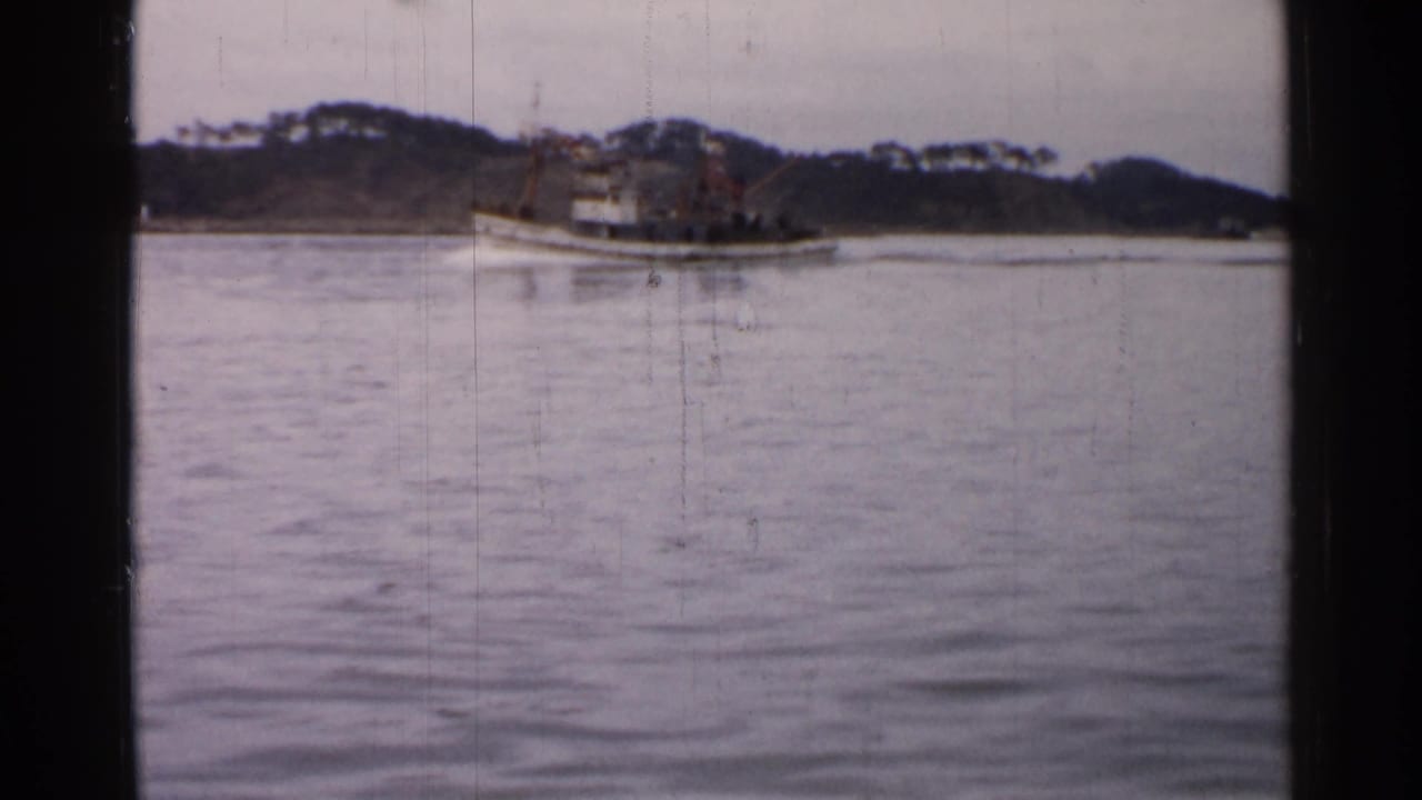 Distant 8mm view of a small boat traveling across calm water in Japan, with a wooded bank and muted sky beyond.