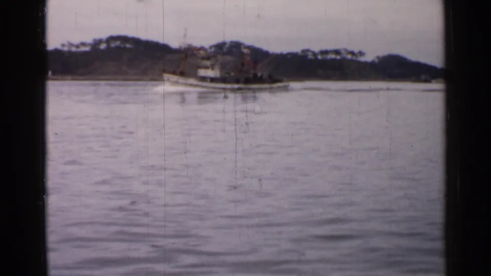 Distant 8mm view of a small boat traveling across calm water in Japan, with a wooded bank and muted sky beyond.