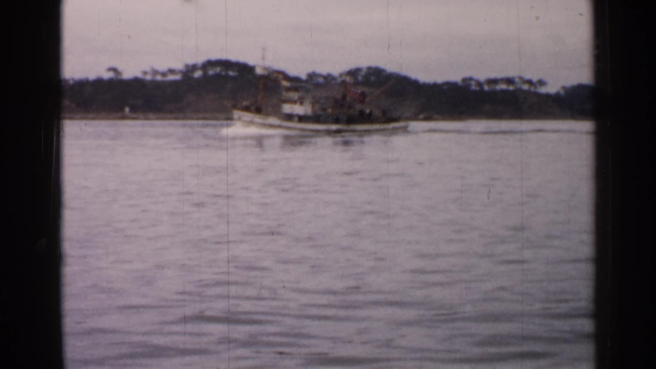 1952 Japan home-movie frame of a small boat on open water, framed by a low wooded shoreline and soft film grain.