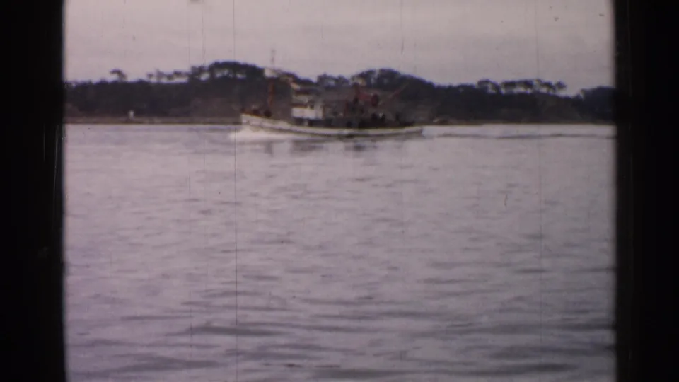 1952 Japan home-movie frame of a small boat on open water, framed by a low wooded shoreline and soft film grain.