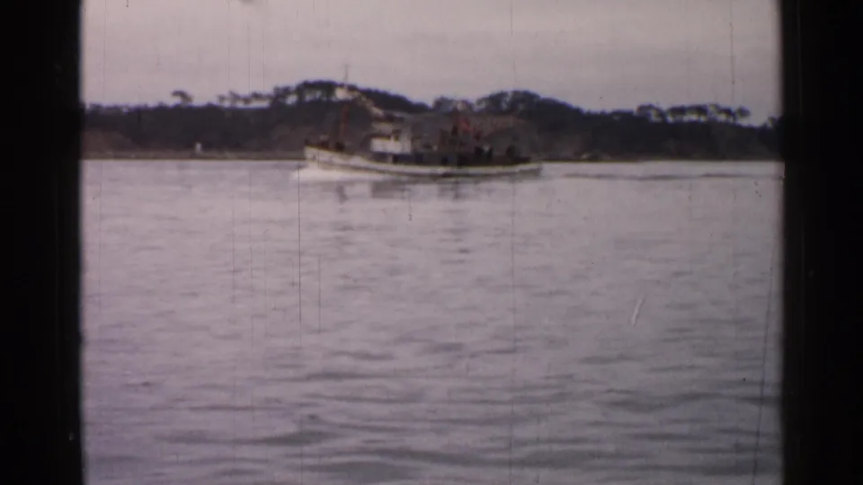 1952 Japan 8mm frame showing a white tugboat on calm water, with small deck figures and a dark wooded shore in the distance.