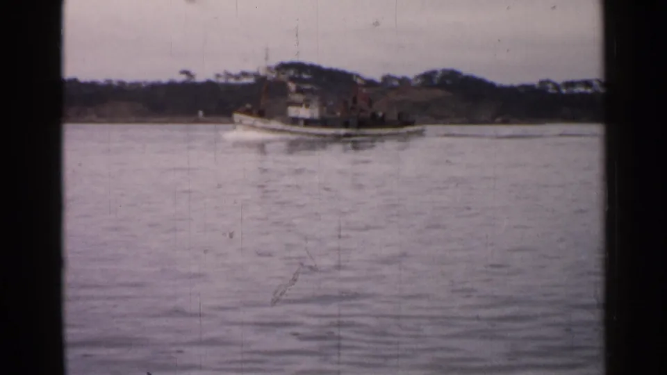 1952 Japan 8mm frame with the white tugboat moving rightward across still water, backed by a dark shoreline and pale sky.